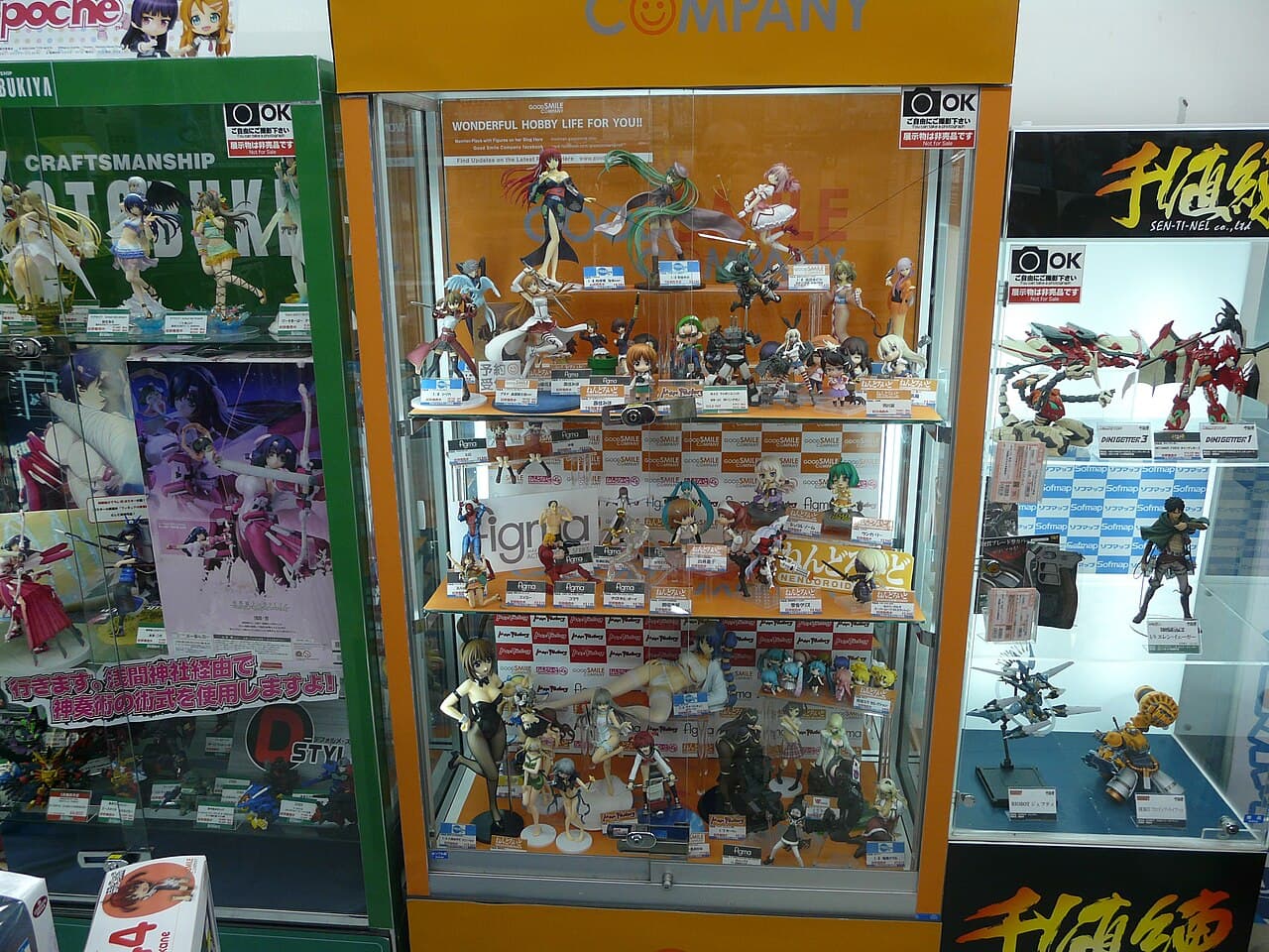 Glass display cabinets packed with Japanese anime figures — including scale figures and figma — at a hobby shop in Akihabara, Tokyo, with Good Smile Company, Kotobukiya and Sen-ti-nel branding and price tags.