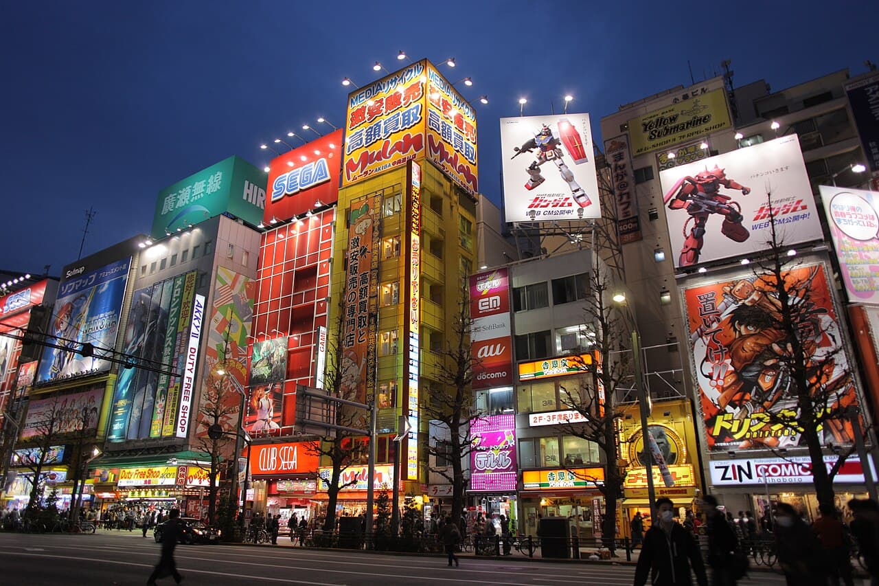 Akihabara Electric Town main street in Tokyo at dusk, lit by towering neon signboards for SEGA arcades, Gundam billboards, and anime, manga and electronics shops, with pedestrians and cyclists on the road below.