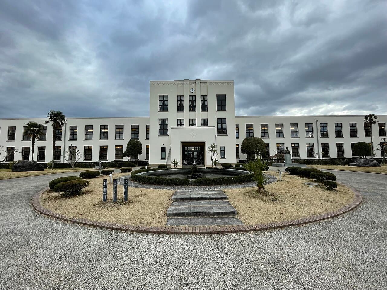The symmetrical white Vories-designed former Toyosato Elementary School building in Toyosato, Shiga, with its central clock tower and long flanking wings, fronted by a circular fountain plaza — the real-life model for the school in the anime K-On!