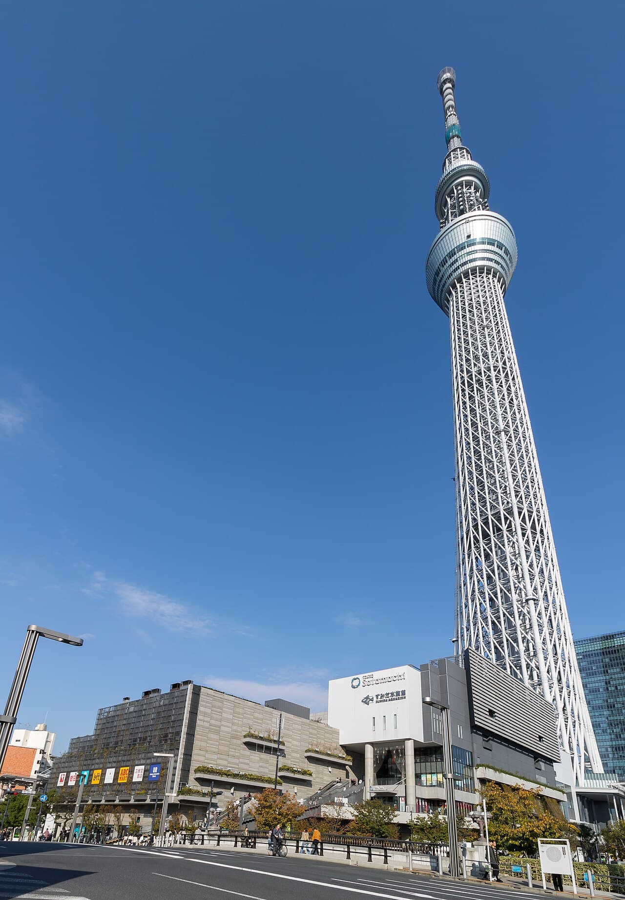 Tokyo Skytree rising above the low Tokyo Solamachi shopping complex under a clear blue sky, seen from street level