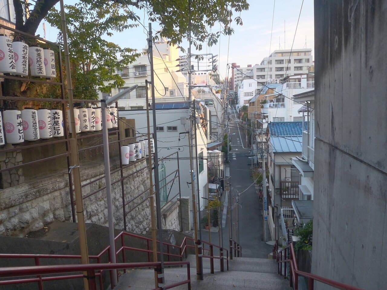High-angle view looking down the red-railed stone staircase of Suga Shrine in Yotsuya, Shinjuku, Tokyo — the stairway made famous by the anime film Your Name — with rows of paper lanterns on the left and a residential street stretching below.