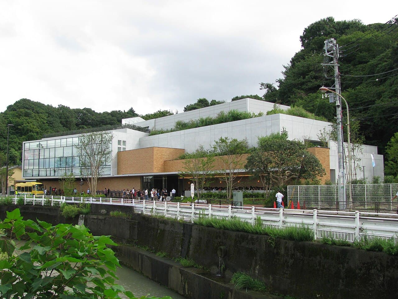 The Fujiko·F·Fujio Museum in Kawasaki: a low building of white panels, brick-coloured walls and a glass corner, with planted roofs. Wooded hills rise behind it, a line of visitors stands along the front, a yellow bus is parked at the left, and a white-railed road and a concrete channel run in the foreground