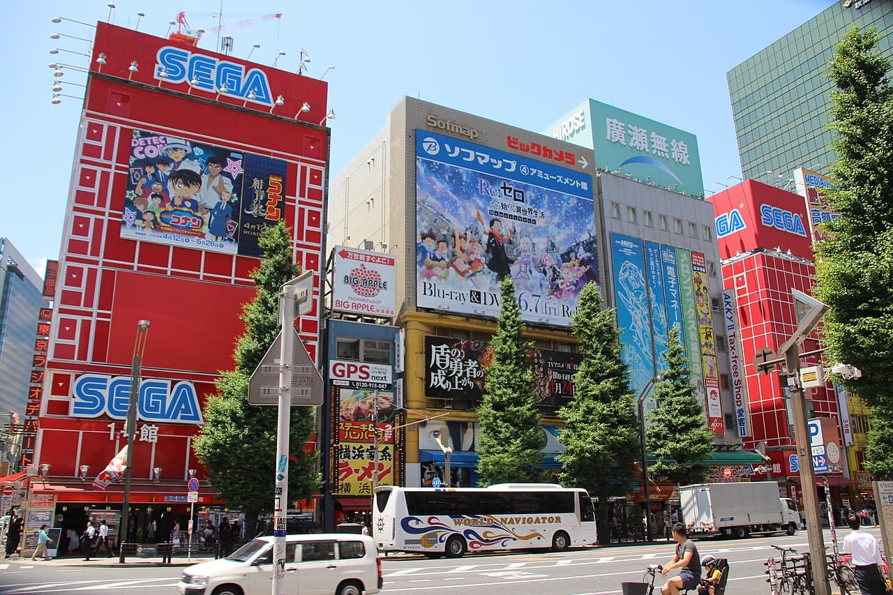 Daytime street view of Akihabara Electric Town in Tokyo, with electronics and anime shop buildings covered in colorful Japanese signs and advertisements.