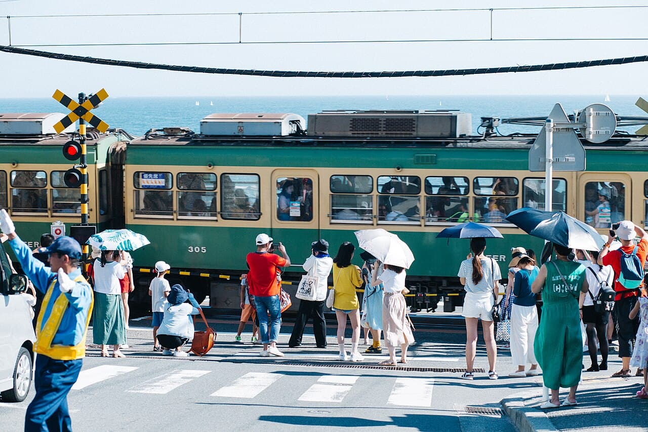 An Enoden (Enoshima Electric Railway) train passing the Kamakura-Kōkō-Mae level crossing in Kamakura — the iconic Slam Dunk opening-scene spot — with tourists gathered by the tracks and the sea beyond.