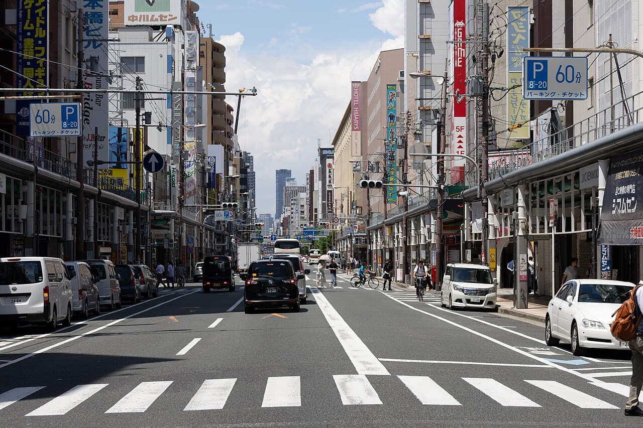 Street view of Den Den Town in Nipponbashi, Osaka, lined with anime, figure and game shop signs under a summer sky.