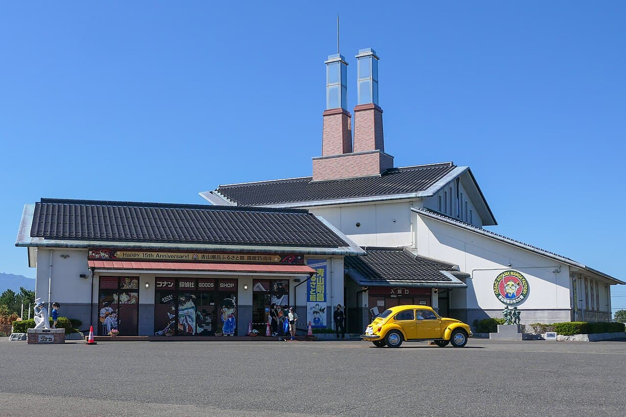 The Gosho Aoyama Manga Factory in Hokuei, Tottori: a white building with dark tiled roofs and twin brick chimney towers, a yellow Volkswagen Beetle parked in front and a 15th-anniversary banner over the shop windows, under a clear blue sky (photo taken September 2022)