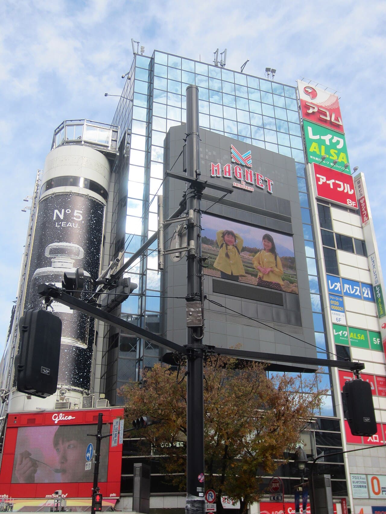 渋谷スクランブル交差点のMAGNET by SHIBUYA109。ガラス張りのビルにMAGNETのロゴと大型ビジョン、左に香水の縦長広告、手前に信号機の柱