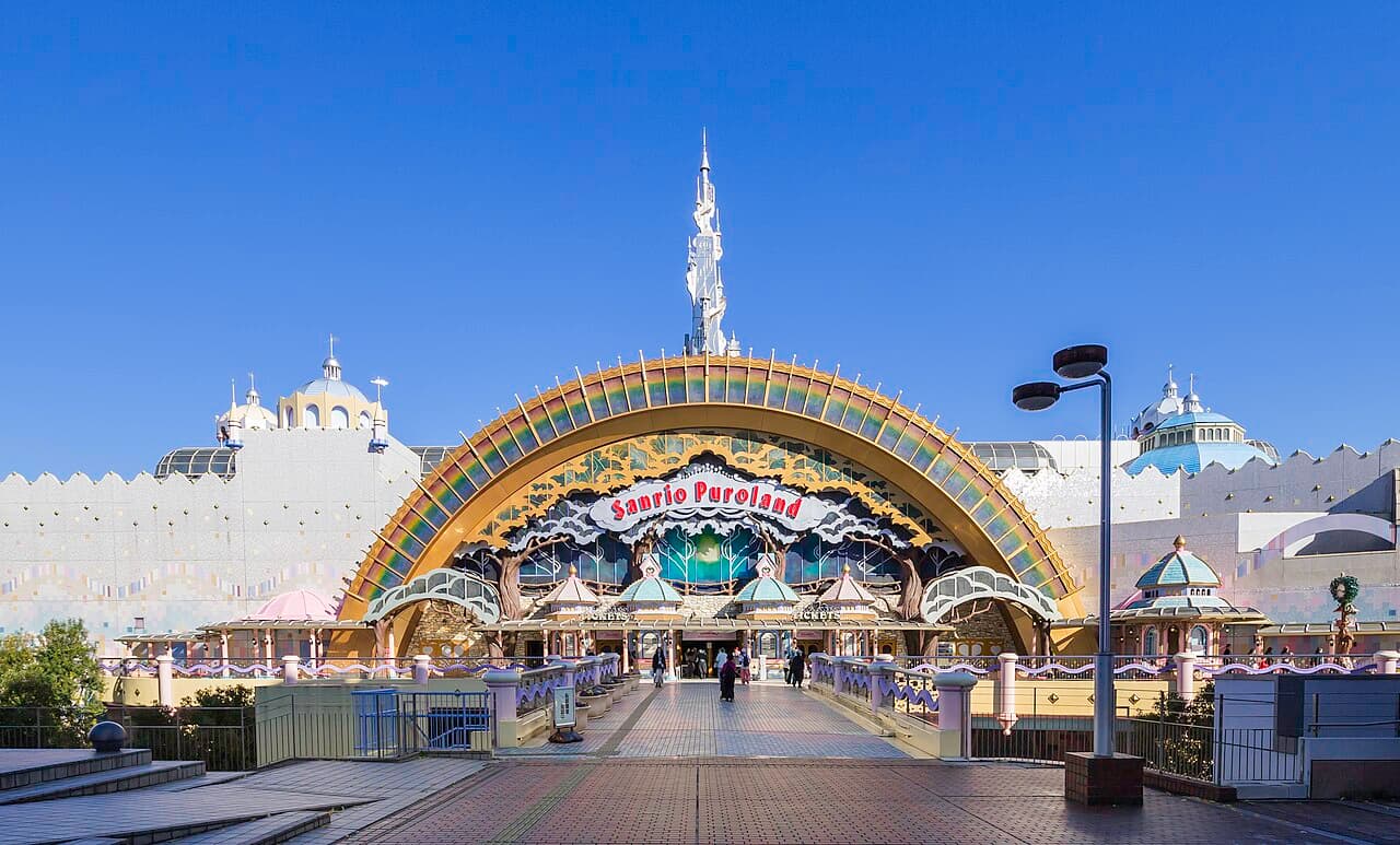 サンリオピューロランドの入口。虹色のアーチに「Sanrio Puroland」の看板と白い尖塔がそびえ、両側に淡い色のドーム屋根の建物、手前に石畳の通路、背景は青空